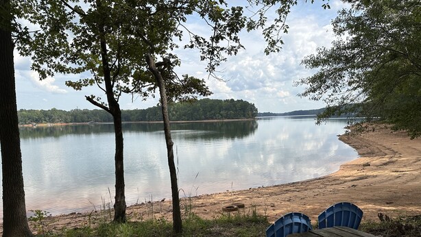 A lakeside view with a wooden picnic table and two blue chairs, surrounded by trees and a calm lake. - Lake Retreat House (Anderson)