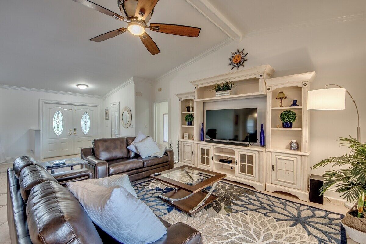 Una sala de estar con un sofá de cuero marrón, una mesa de centro de vidrio, un mueble blanco para televisión con pantalla plana y un ventilador de techo.