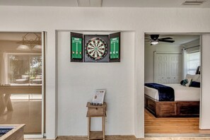 A room with a dartboard, a small wooden side table with a magazine, and a bed with a blue blanket.