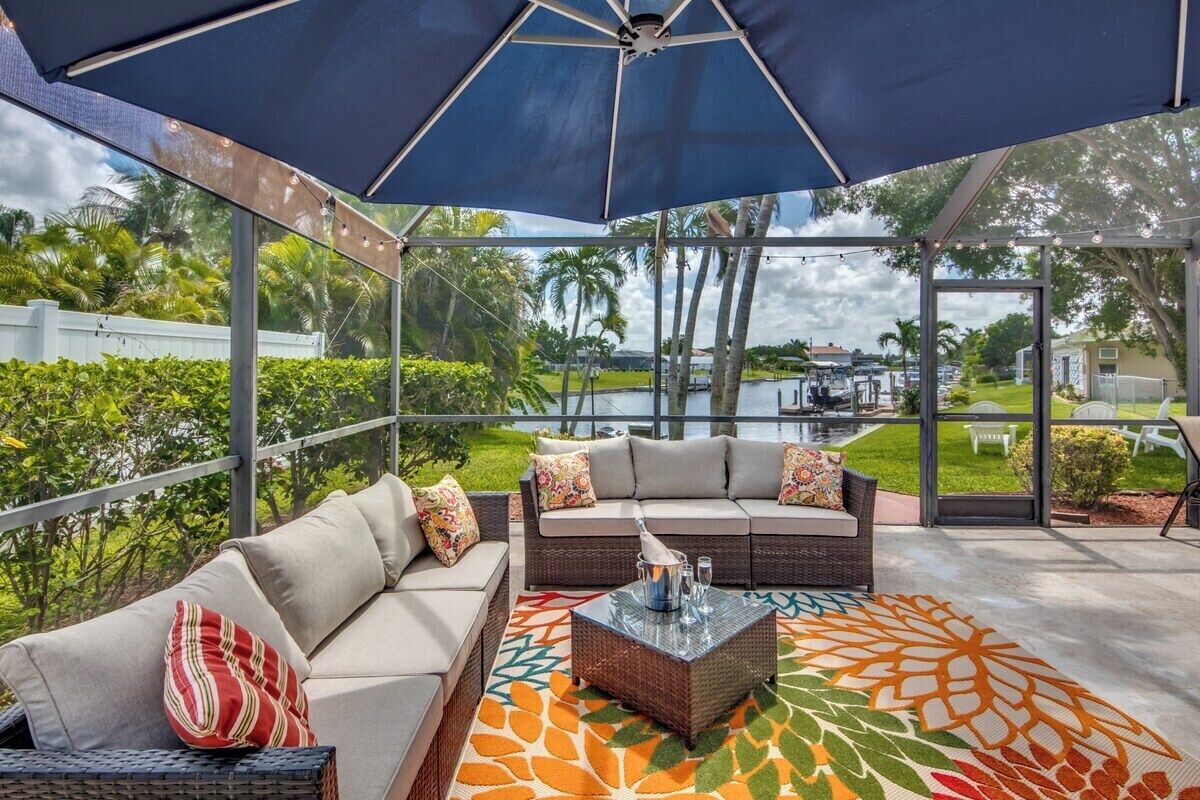 An outdoor patio with a sofa and a coffee table, a large umbrella, and a view of a body of water and greenery.
