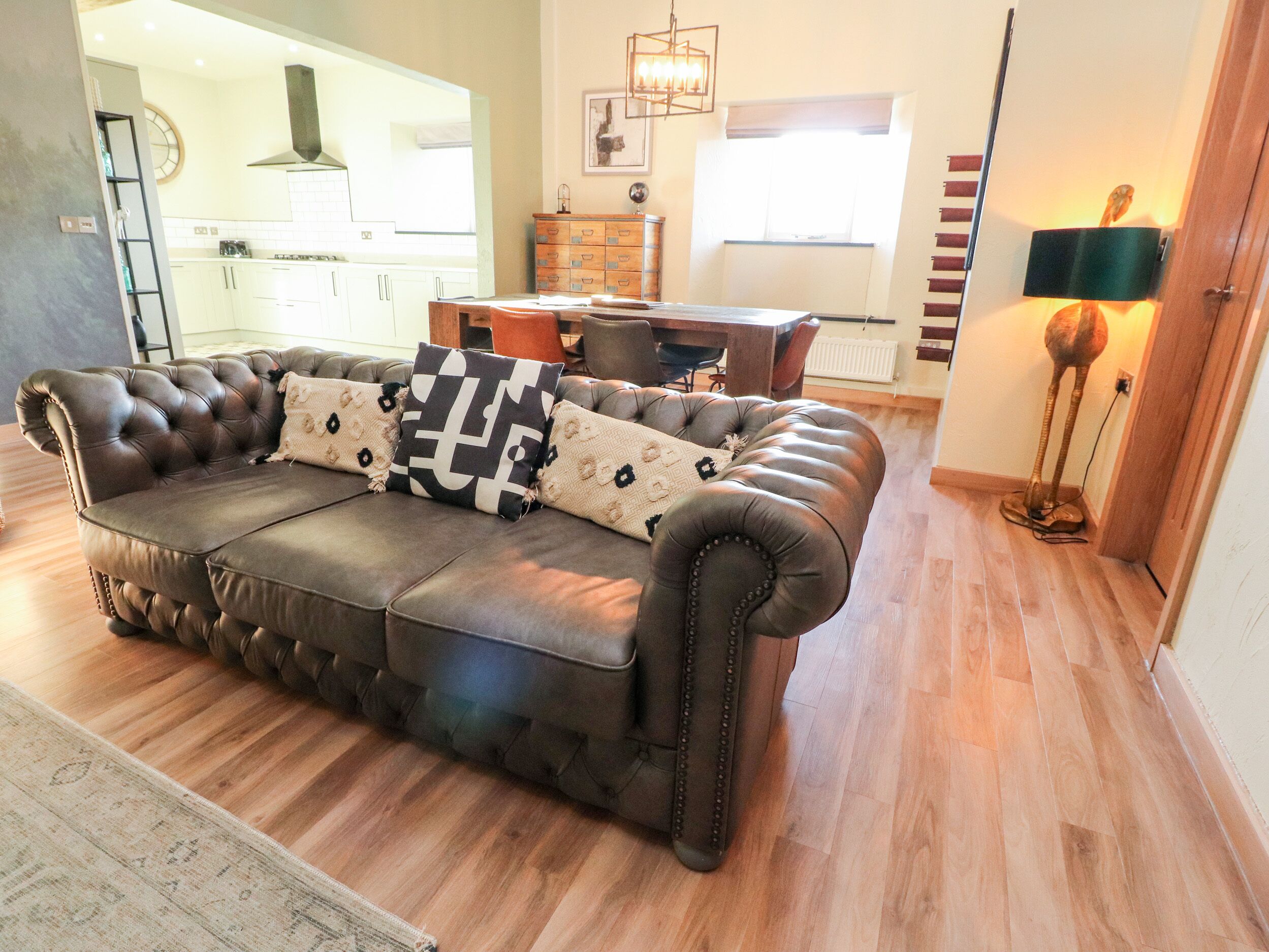 A living room with a dark brown leather sofa, patterned cushions, a wooden coffee table, and a dining area in the background.