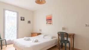 A neatly arranged bedroom with a bed, a desk, a chair, a nightstand, and a door leading to a balcony. - LES AJONCS - 300m de la plage - Parking (Batz-sur-Mer)