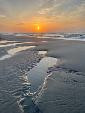 Uma praia com padrões de areia molhada e um pôr do sol ao fundo.