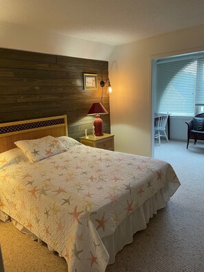 A bedroom with a large bed, a nightstand with a lamp, a wooden headboard, and a window with blinds. - Steps to the beach, quiet area of beach. (Surfside Beach)