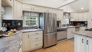Una cocina con armarios blancos, un refrigerador de acero inoxidable, una ventana con vistas y una encimera con tabla de cortar y uvas.