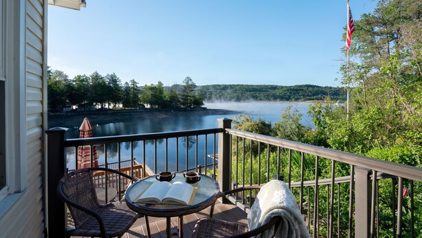 Un balcón con vista a un lago, una bandera y muebles de exterior, incluyendo una mesa con un libro y dos tazas.