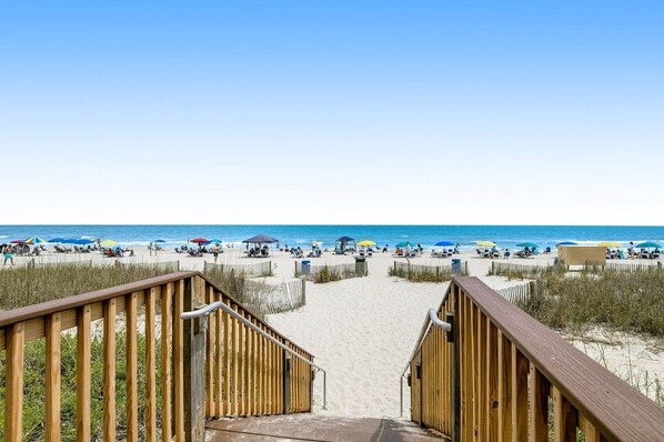 A wooden staircase leading to a sandy beach with umbrellas and beachgoers.