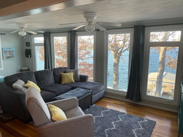 A living room with a dark gray sectional sofa, a light gray armchair, and a gray patterned rug. There are two ceiling fans, large windows with curtains, and a view of a body of water and trees.