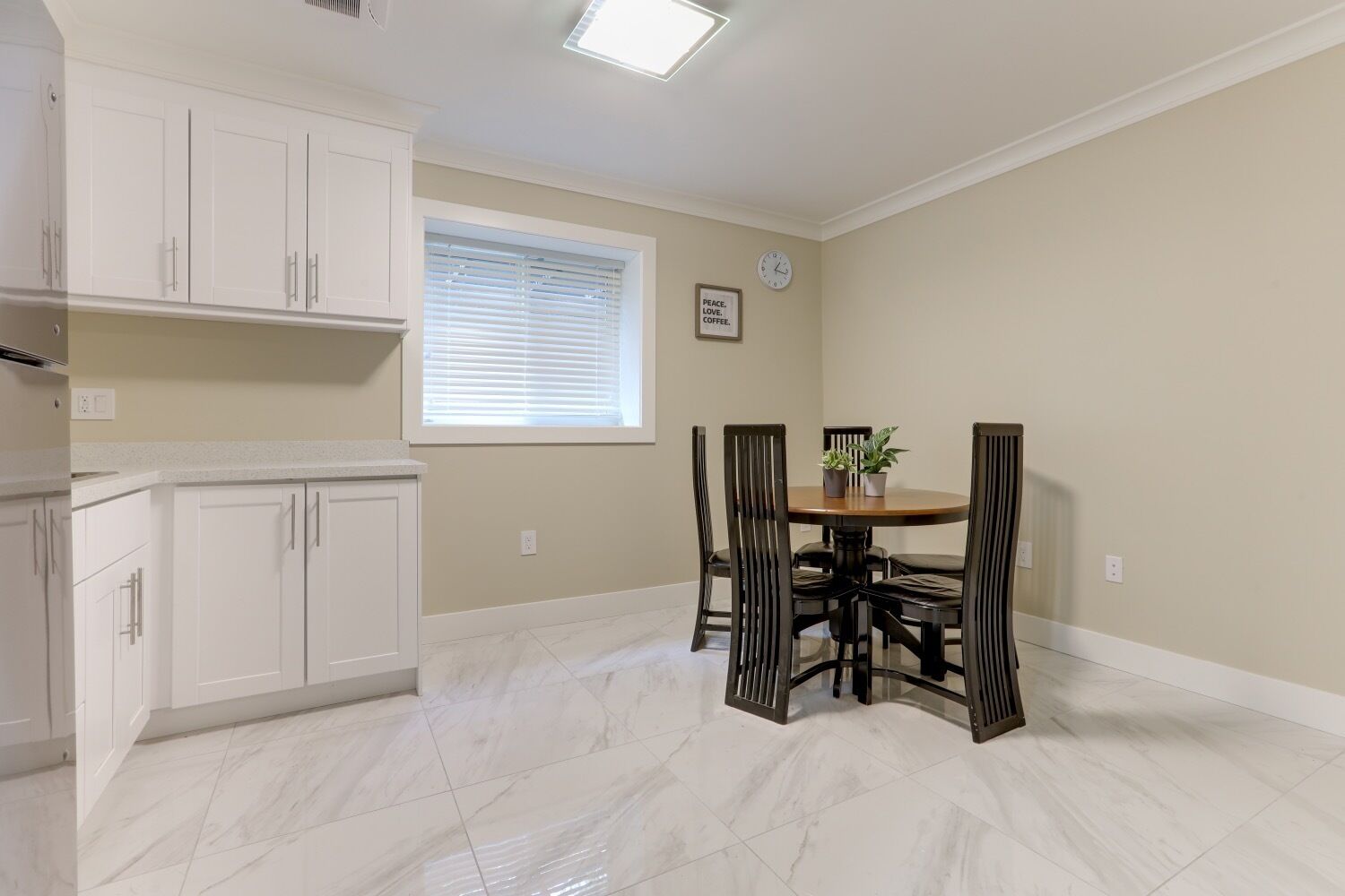 Un comedor con una mesa redonda y sillas, una ventana con persianas y muebles de cocina blancos.