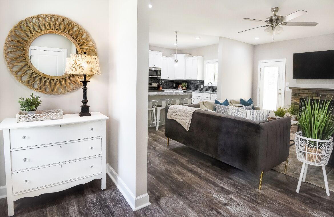 Un salon moderne avec un canapé foncé, une commode blanche et un miroir rond doté d’un cadre doré.