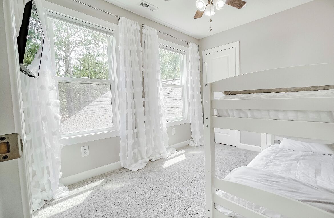 Une chambre avec un lit superposé, de grandes fenêtres avec des rideaux légers, un ventilateur de plafond et une porte.