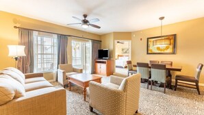 A hotel room with a sofa, armchairs, a dining table with chairs, a TV, and a ceiling fan.