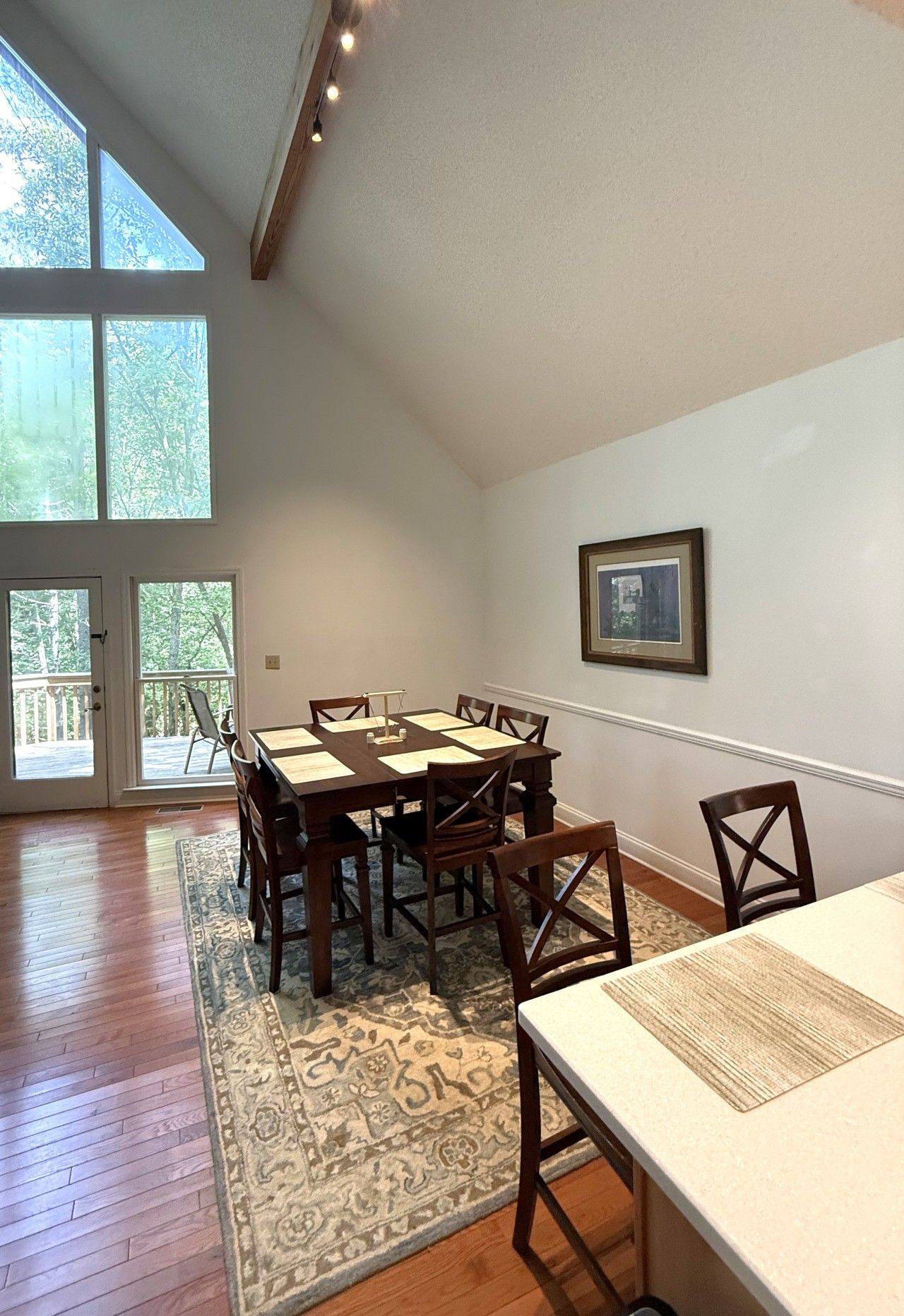 A dining area with a wooden table and chairs, a rug, and a large window with a view of trees.