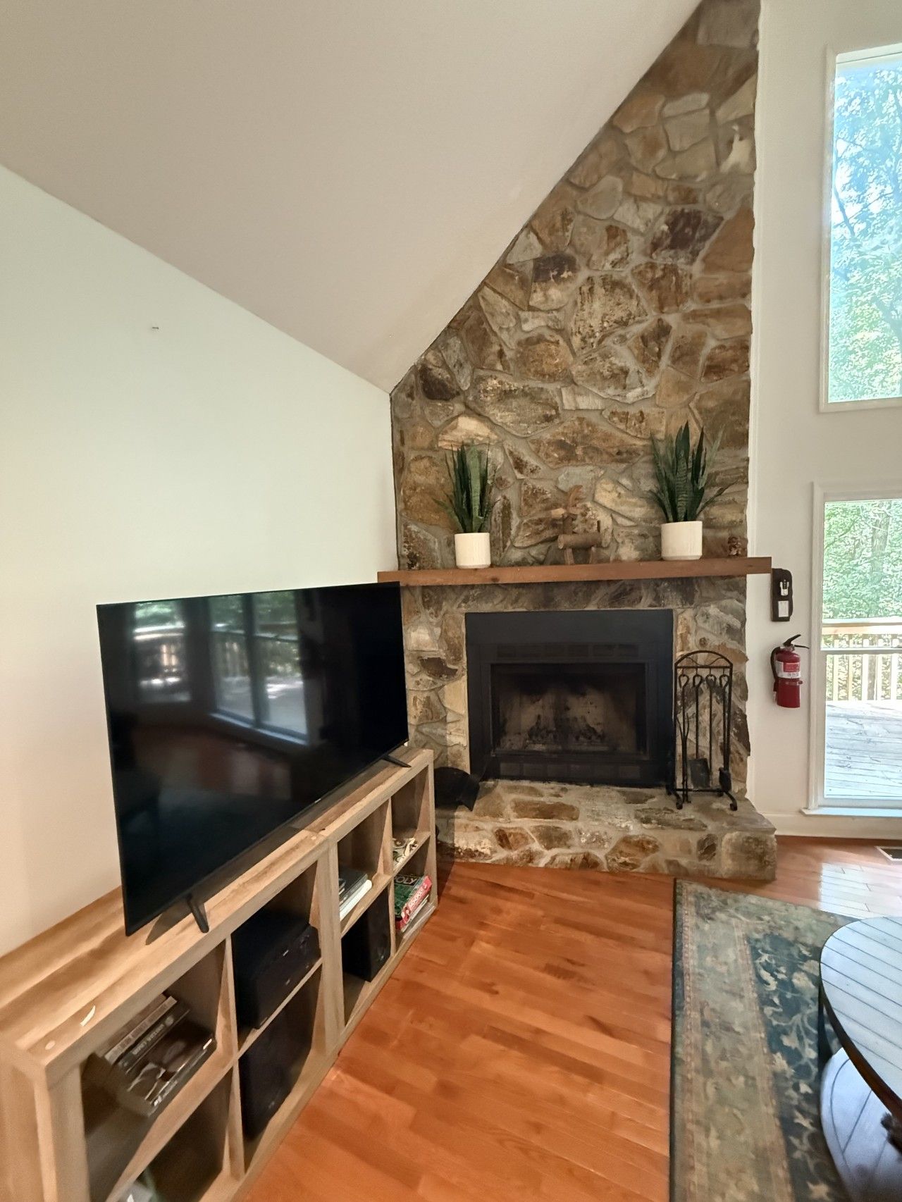A living room with a stone fireplace, a flat-screen TV on a wooden stand, and a fire extinguisher on the wall.
