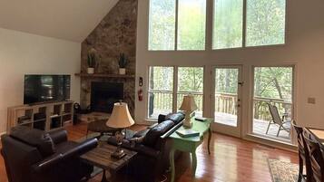 Una sala de estar con chimenea de piedra, televisor, sofás de cuero y ventanas grandes con vistas a árboles.