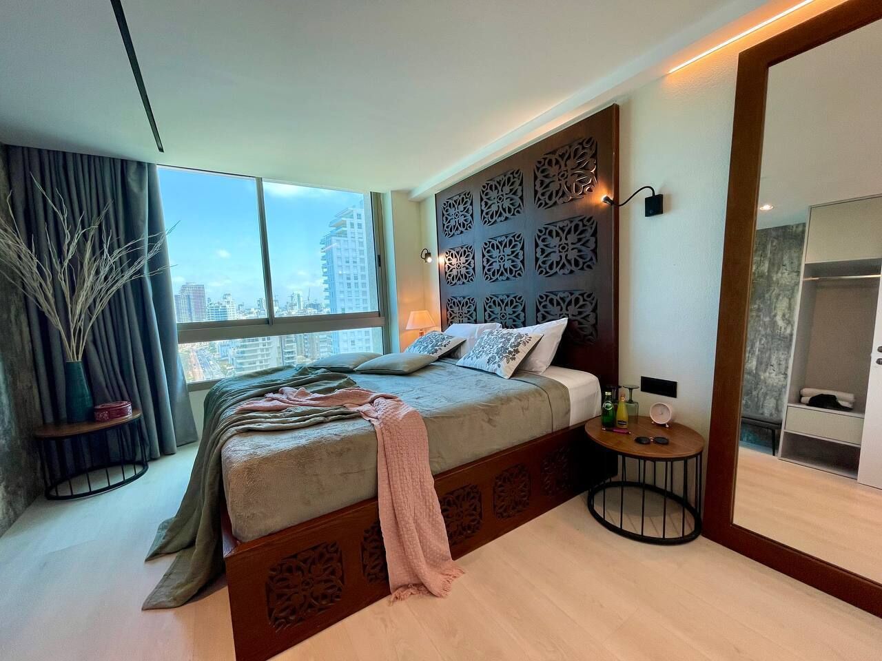 Un dormitorio moderno con una cama grande, vistas a la ciudad, un cabecero decorativo y una mesita de noche con una lámpara.