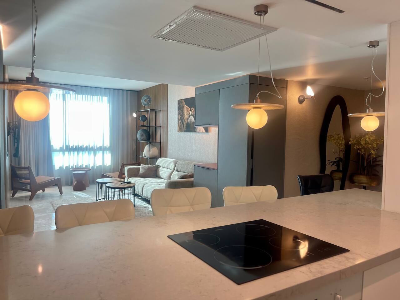 Una sala de estar moderna con comedor, una cocina integrada con placa de inducción negra y un ventanal con cortinas.
