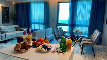 A modern living room with a dining area, a sofa, a glass table with food and drinks, and a view of the sea through large windows.