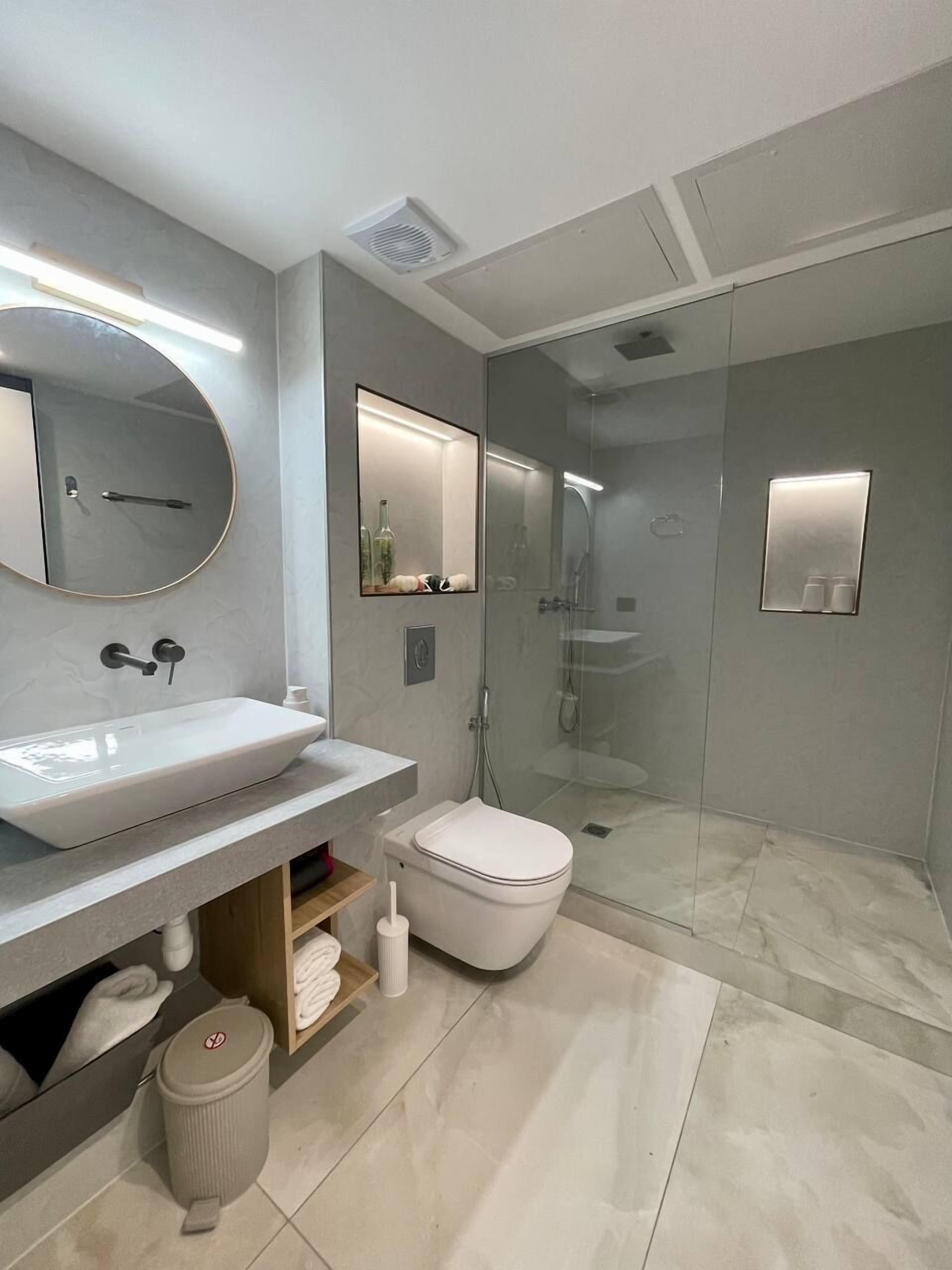 Un baño moderno con un espejo ovalado grande, un espejo rectangular, un lavabo blanco, un inodoro y una zona de ducha con mampara de vidrio.