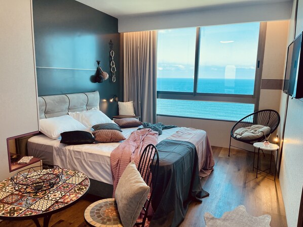 Un dormitorio moderno con una cama grande, una mesita redonda pequeña, una silla y vistas al mar a través de amplias ventanas.