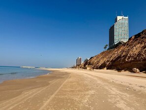 Una playa con cielo despejado, orilla arenosa y un edificio alto al fondo.