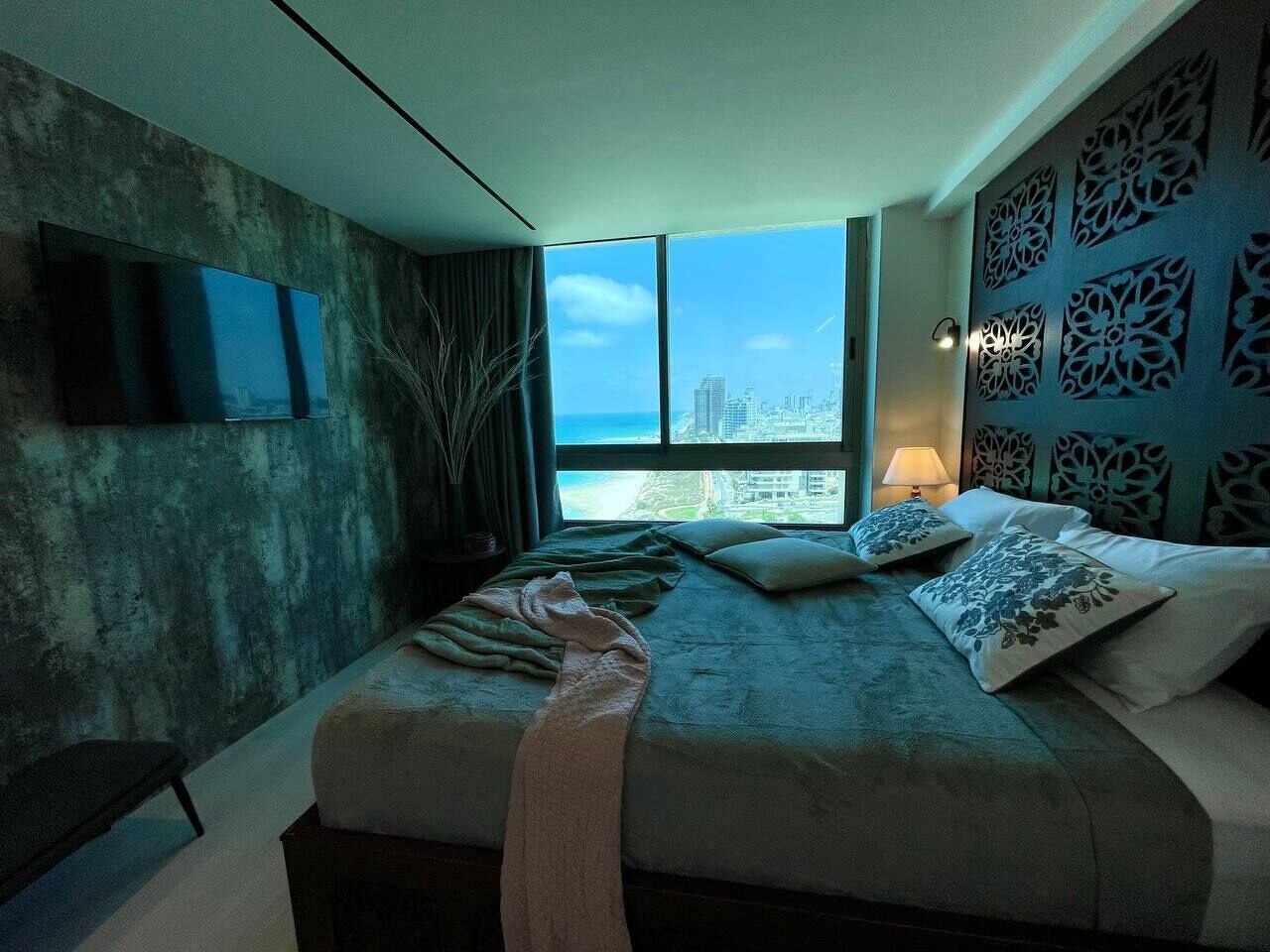 Ein modernes Schlafzimmer mit einem großen Bett, einem an der Wand befestigten Fernseher und Blick auf die Stadt durch große Fenster.