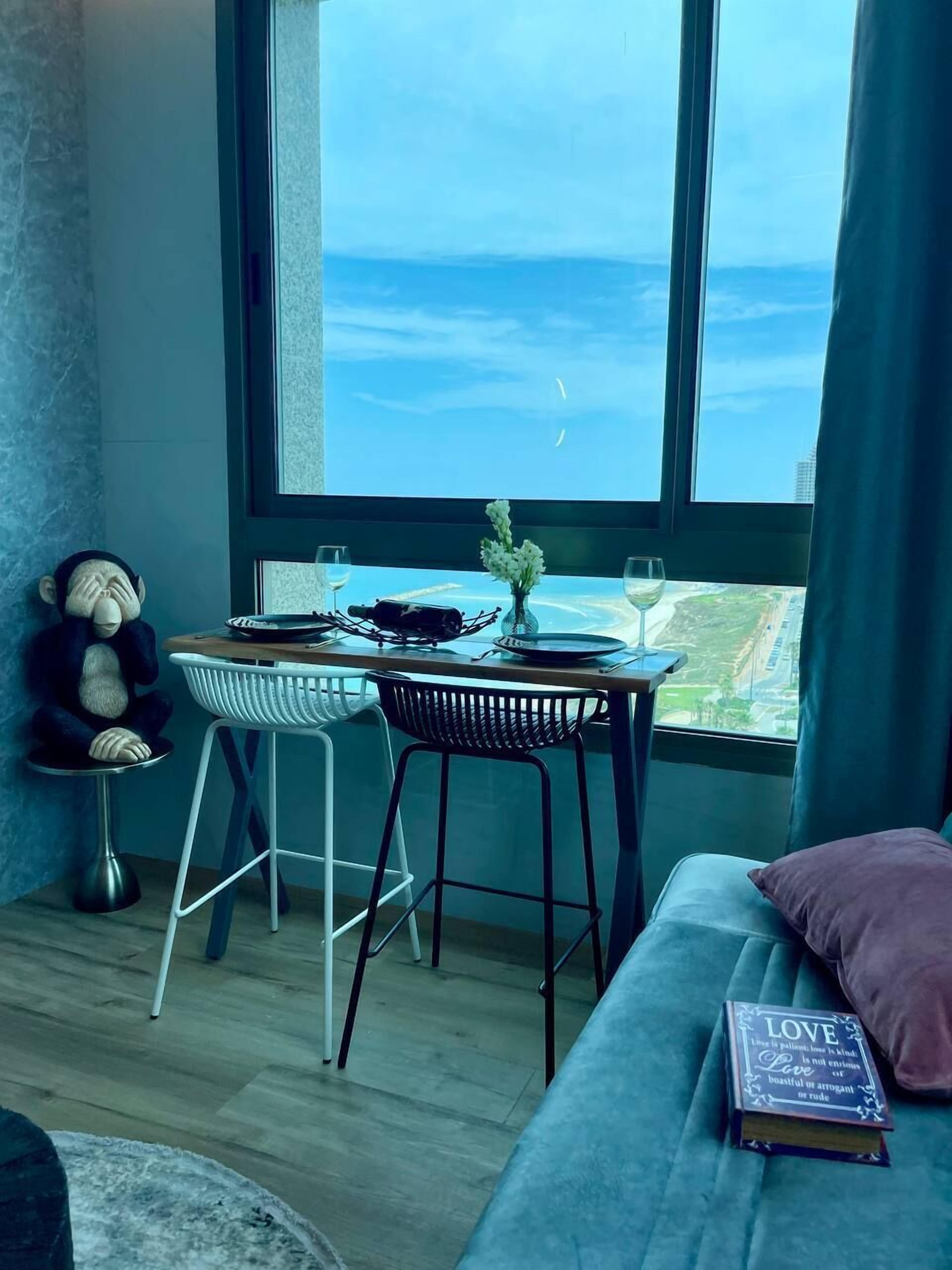 Een gezellige kamer met een groot raam, een klein tafeltje met een vaas, een teddybeer en een boek op een bank.