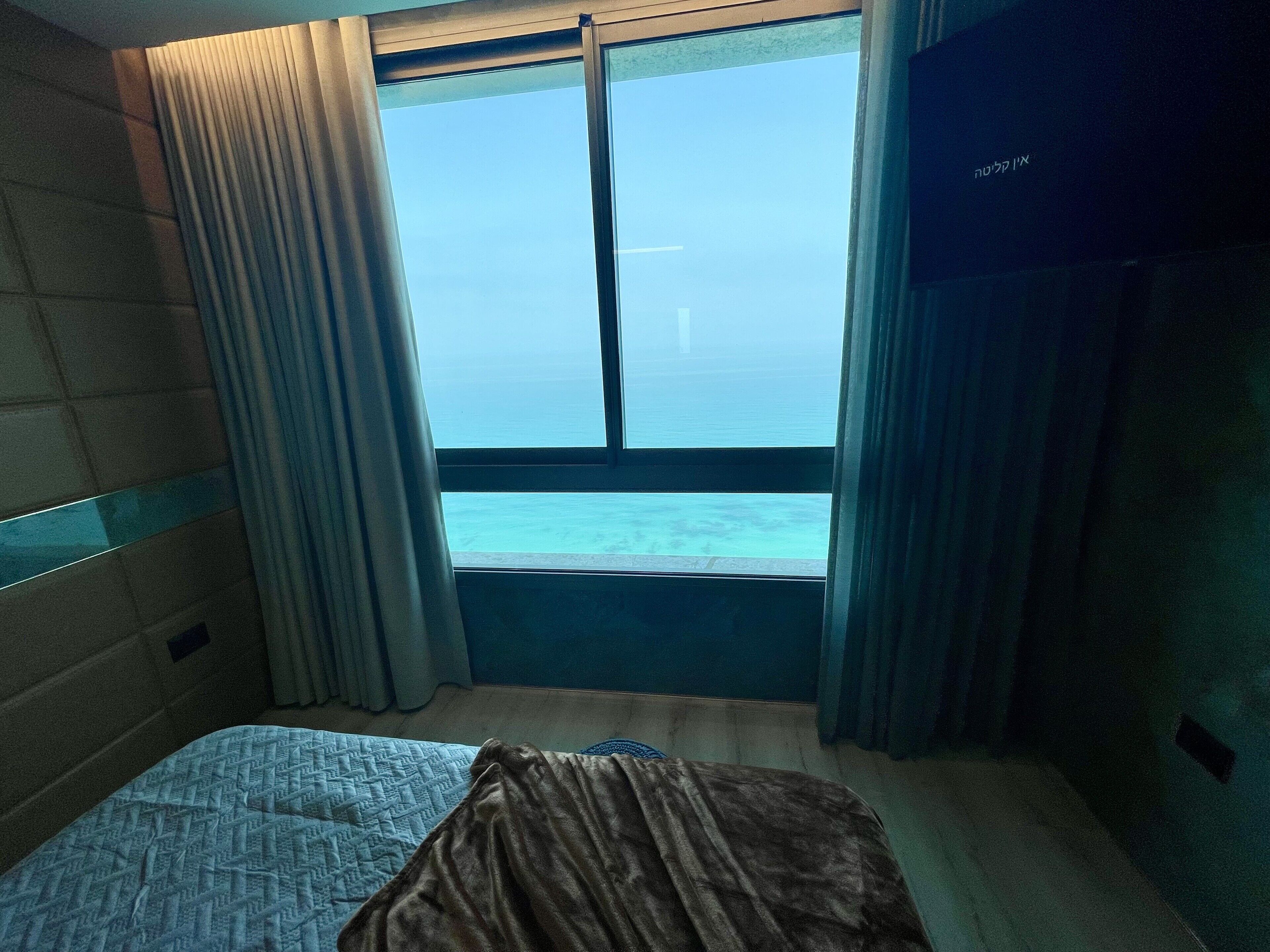 Una habitación de hotel con cama, un ventanal con cortinas y vista al mar.