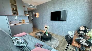Un espacio habitable moderno con un sofá gris, un comedor con mesa de madera y una cocina con gabinetes grises y un televisor de pantalla plana montado en la pared.