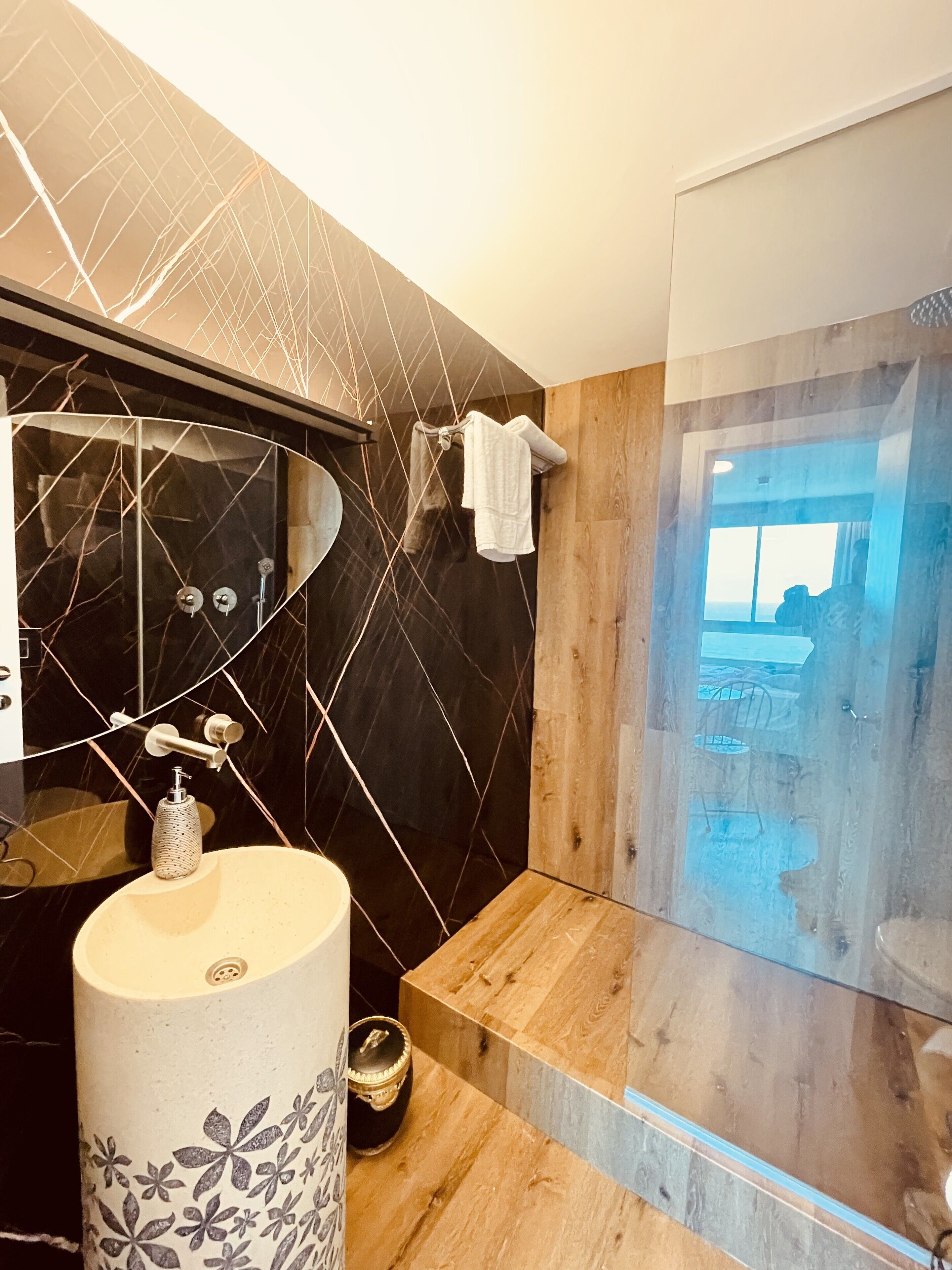 Ein modernes Badezimmer mit Waschtisch aus schwarzem Marmor, einer Duschkabine aus Holz und einer Glastür, die auf einen Balkon führt.