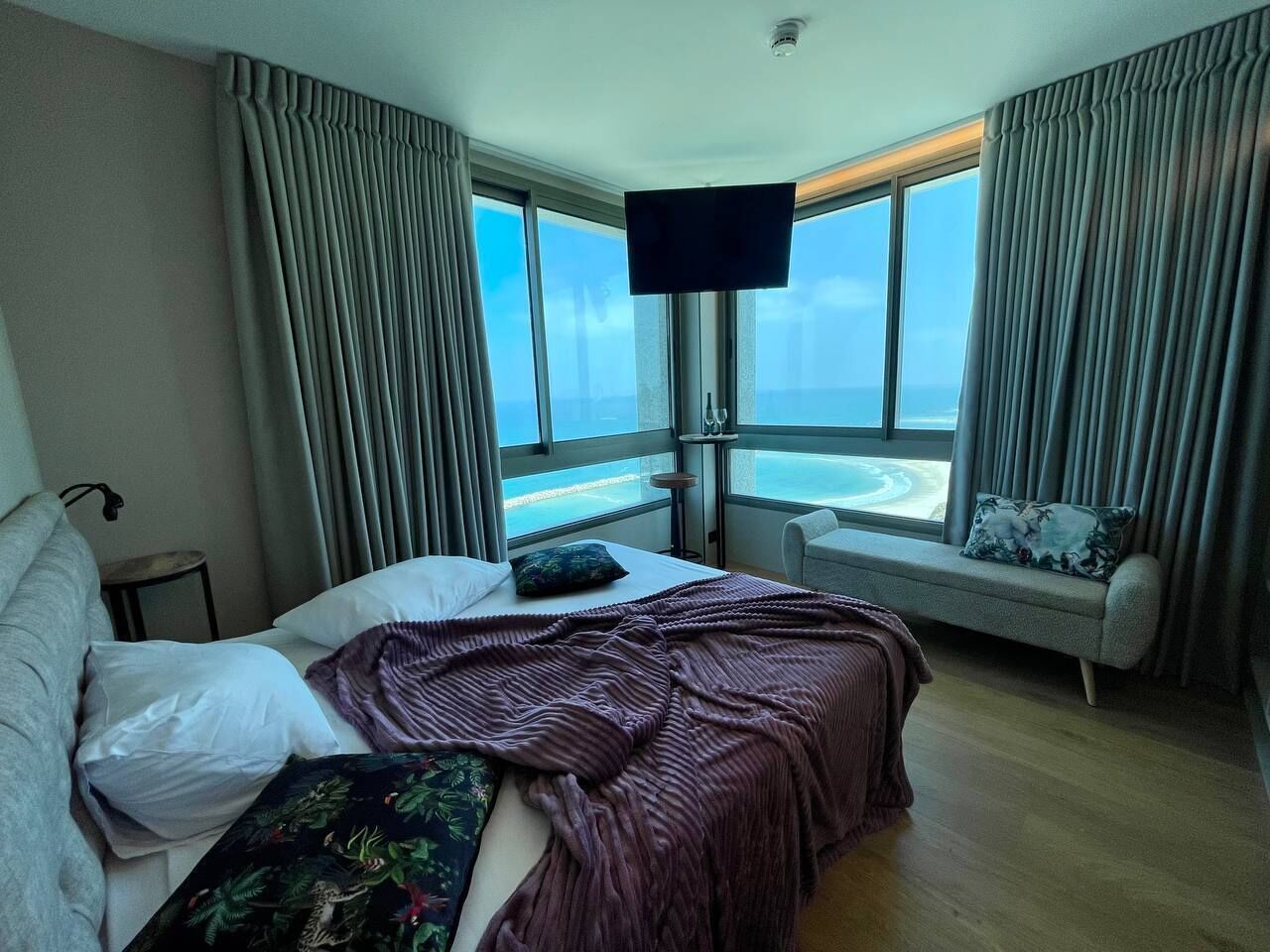 A hotel room with a bed, a window with curtains, a TV mounted on the wall, and a view of the sea.