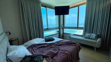 A hotel room with a bed, a window with curtains, a TV mounted on the wall, and a view of the sea.
