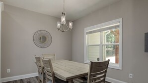 A dining room with a wooden table and chairs, a chandelier, a wall-mounted mirror, and a window with a view of trees. - Serenity on Pinehurst No 6! <3.5 miles to the Village of Pinehurst! Sleeps 10! (Pine Hurst)