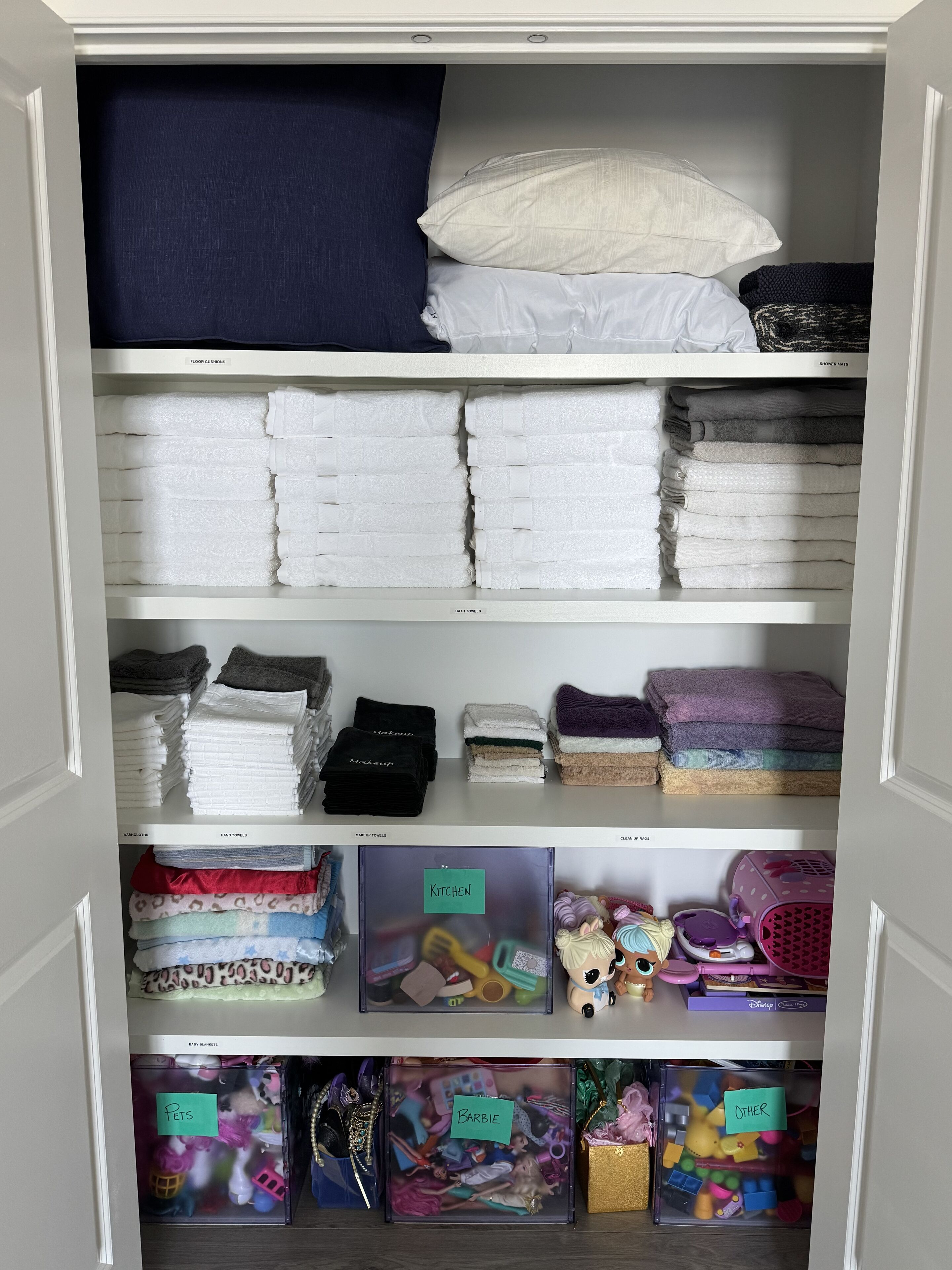A closet with shelves labeled for different types of towels and blankets, including 'floor cushions,' 'shower mats,' 'hand towels,' 'makeup towels,' and 'cleaning mats.' There are also bins labeled 'pets,' 'barbie,' and 'other.'