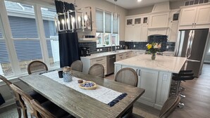 A modern kitchen with white cabinets, a stainless steel refrigerator, and a dining area with a wooden table and chairs. - SUNSET RETREAT-Luxury 4BR, 4.5BA waterfront, boat lift, best sunset on the lake! (Russells Point)