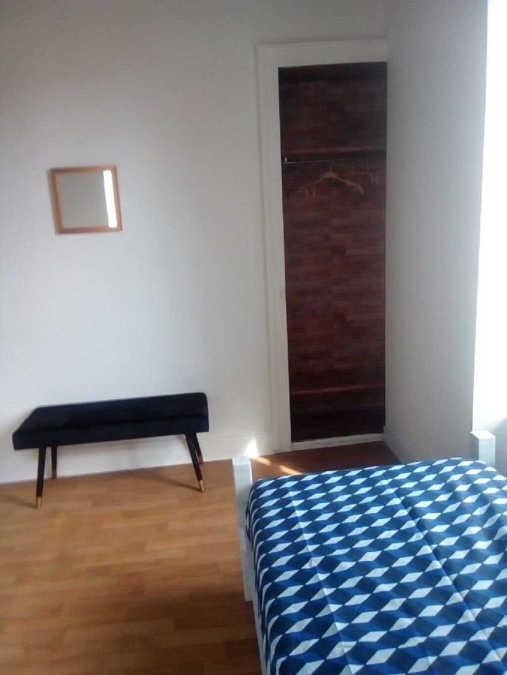Een slaapkamer met een houten vloer, een bed met een blauw-wit geruit dekbed, een zwarte bankje, een witte nachtkastje en een houten deur.