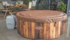 Un bain à remous en bois, doté d’un couvercle marron et portant la marque « SALUSPA HELSINKI », installé à l’extérieur, avec un sol carrelé et une table entourée de chaises en arrière-plan.