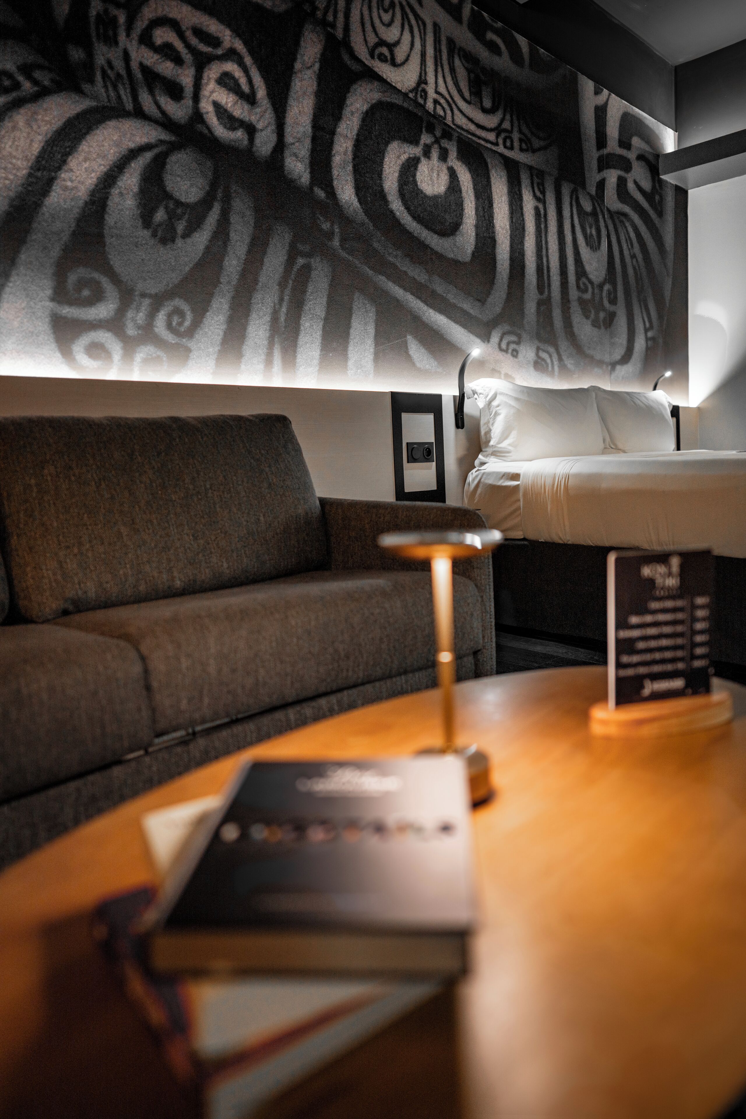 Quarto de hotel com cama, sofá, mesa com abajur e parede com padrão decorativo.