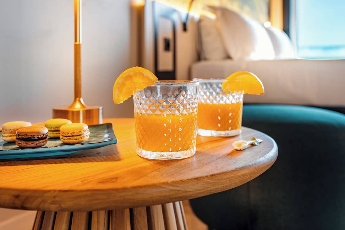 Een houten tafel met twee glaasjes sinaasappelsap, een lamp en macarons.