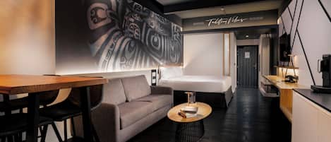A modern hotel room with a sofa, a coffee table, a dining area, and a large abstract wall art piece.