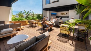 Een buitenterras met rieten meubels, houten tafels en stoelen. Een groot scherm is aan de muur bevestigd en het gebied is omringd door groen.