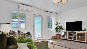 Ein modernes Wohnzimmer mit einer Couch, einem Couchtisch mit Pflanze, einem Fernseher auf einem Holzgestell und großen Fenstern mit Jalousien.