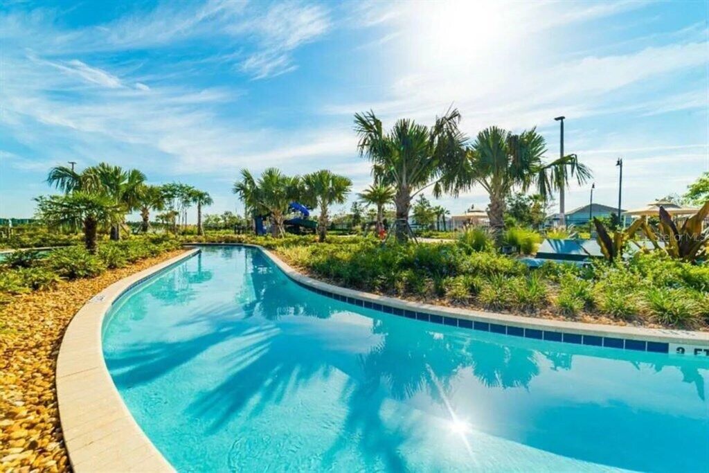 A curved swimming pool surrounded by palm trees and greenery, with a clear blue sky above.