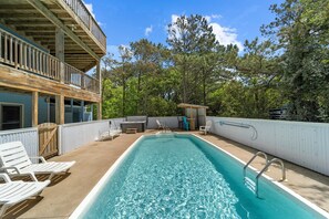 A rectangular pool with a wooden deck, white lounge chairs, and a small wooden structure.