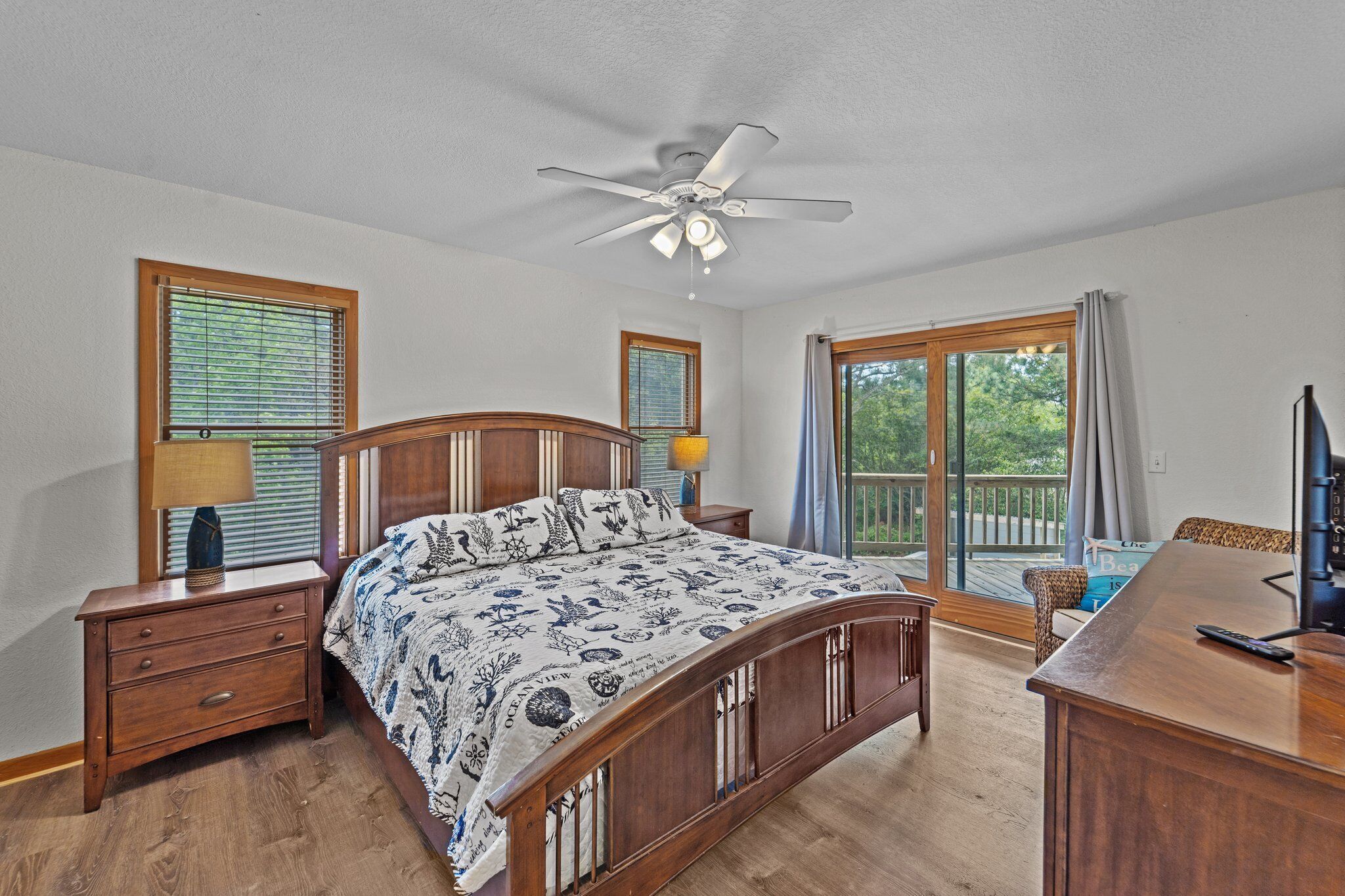 A bedroom with a wooden bed, a ceiling fan, a nightstand, a dresser, and a view of the outdoors through large windows.