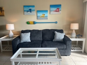 A living room with a dark grey sofa, two white side tables with lamps, a white coffee table, and three beach-themed paintings on the wall.