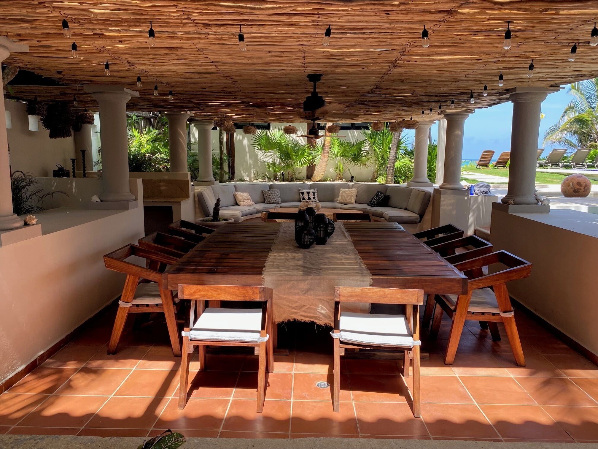 A covered outdoor dining area with a long wooden table and chairs, a sofa, and a ceiling with hanging light fixtures.