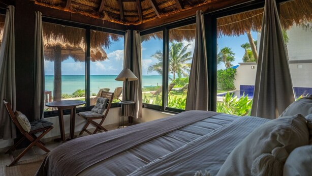 A bedroom with a large bed, a small table, and two chairs. It has a view of the ocean and palm trees. - BEACHFRONT LUXURY VILLA ON A PRIVATE BAY (Tulum)