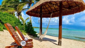 A beach scene with a wooden lounge chair, a hammock, and a thatched umbrella. - BEACHFRONT LUXURY VILLA ON A PRIVATE BAY (Tulum)
