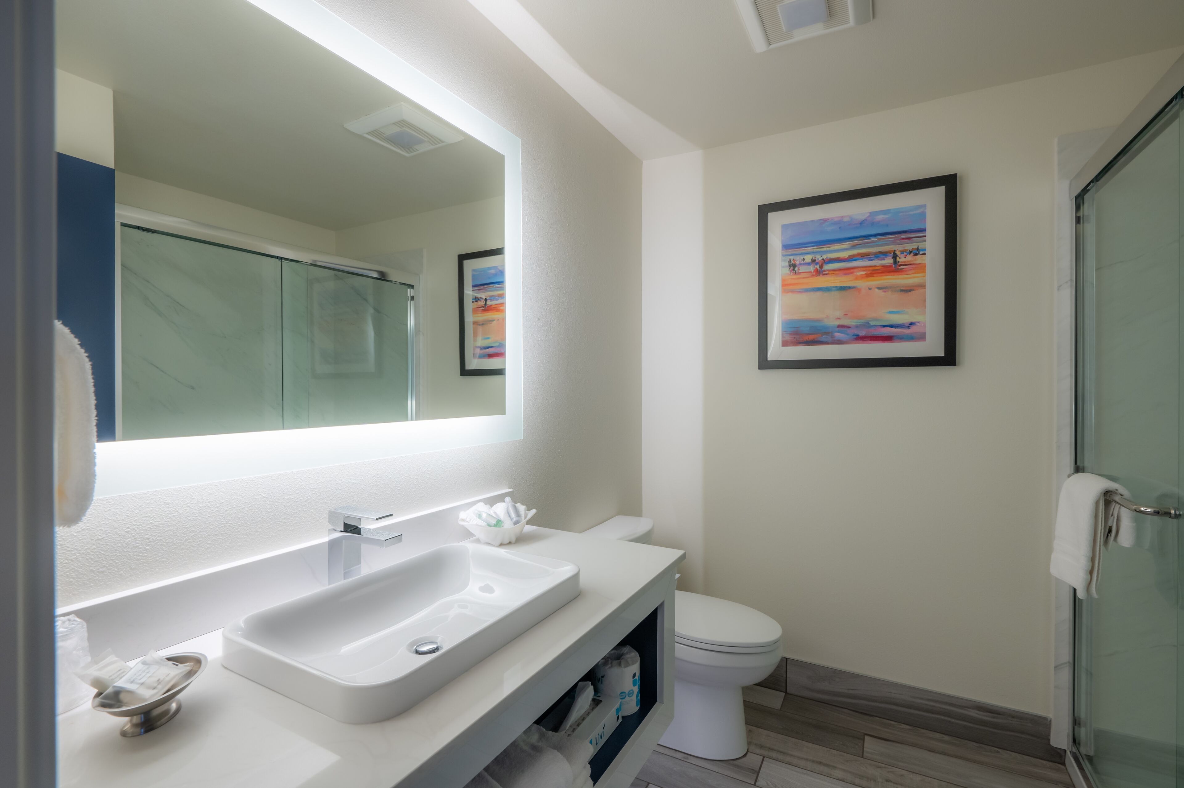 A modern bathroom with a white sink, mirror, and toilet. There is a framed picture on the wall and a towel rack.